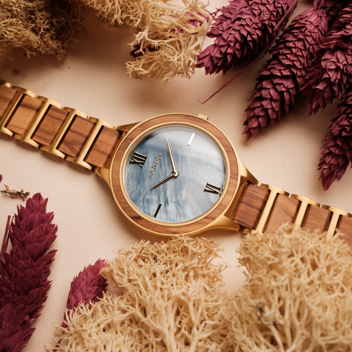 Orologio "Capri" con finitura in legno naturale ed elegante quadrante nacre, circondato da fogliame essiccato.