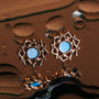 "Composition Earrings" earrings feature intricate rose gold with blue nacre accents, set against a reflective surface.