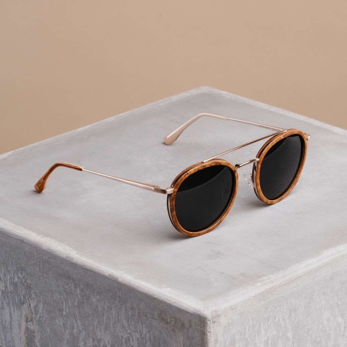 Gli occhiali da sole "Incantesimo" caratterizzati da una cornice in legno di cannella e da accenti in oro opaco su uno sfondo minimalista in cemento.