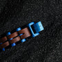 "Kanon" watch strap, walnut wood with blue accents, laid on textured black surface.