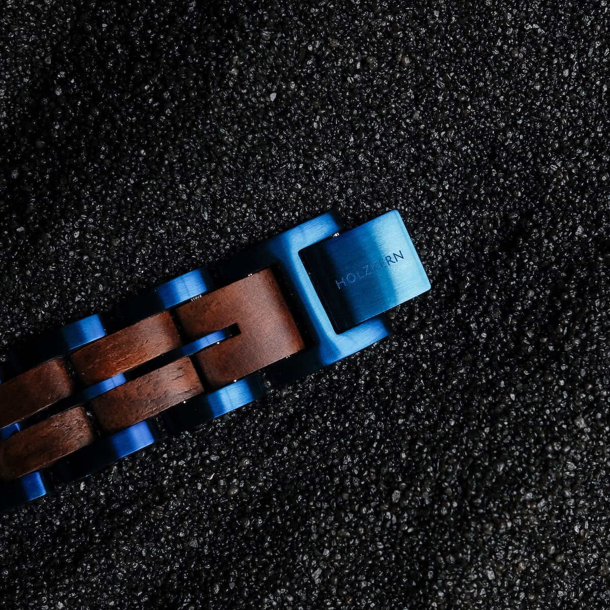 "Kanon" watch strap, walnut wood with blue accents, laid on textured black surface.