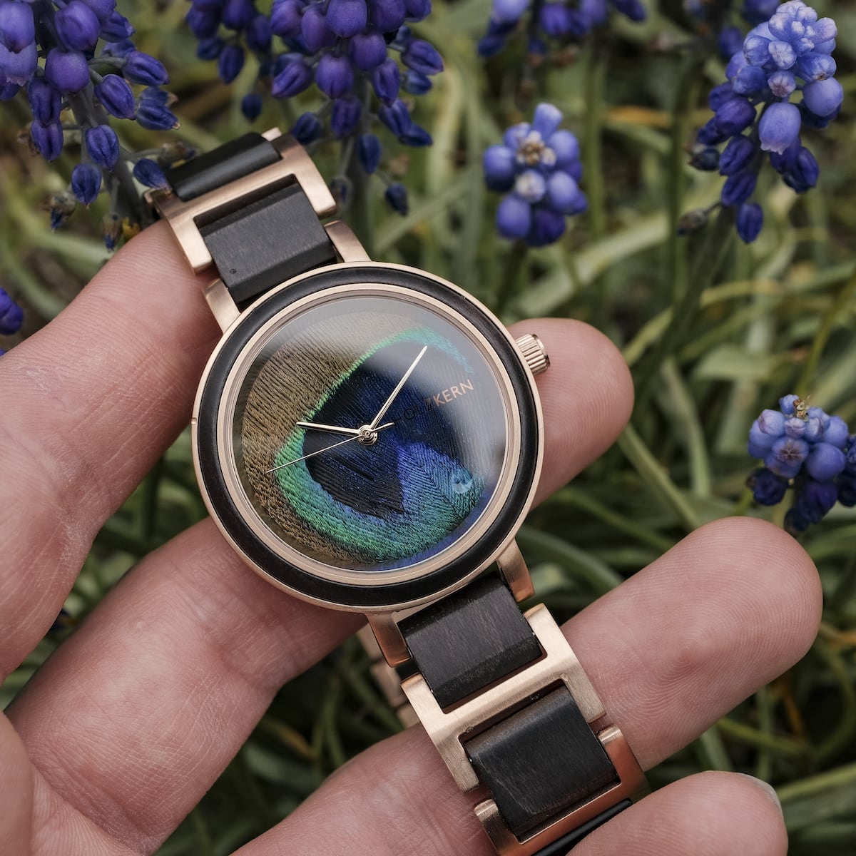 "The Fearless" orologio con un sorprendente quadrante in piuma di pavone e elementi acero, ambientato su uno sfondo di fiori vivaci.
