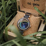 "The Fearless" orologio con una banda di acero e pavone faccia di piuma, ambientato in una elegante scatola di legno circondata da erba.