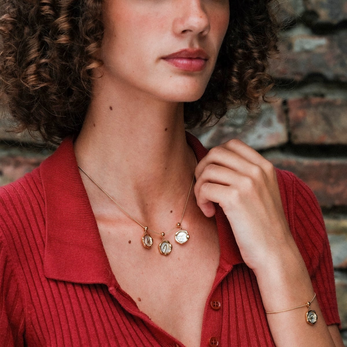 "Letter Charm A-Z", collana di fascino letterale in nacre e oro, indossata da una persona in una parte superiore rossa su uno sfondo di mattoni.