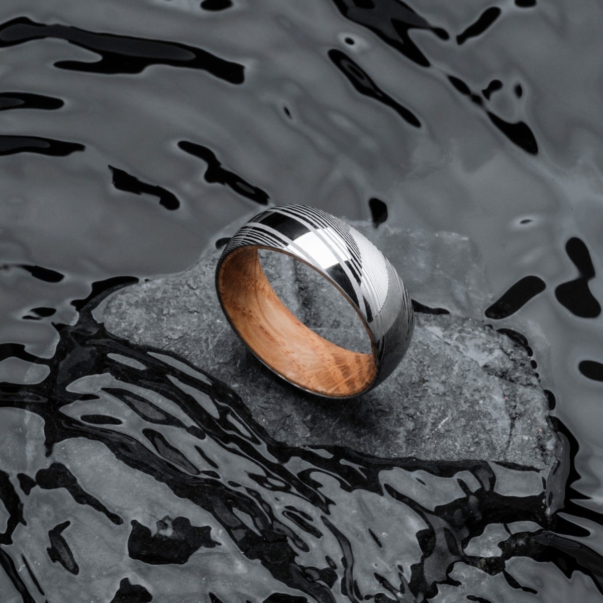 "Sumo Ring", a sleek ring crafted from oak and Damascus steel, elegantly displayed on a stone in a water-inspired setting.