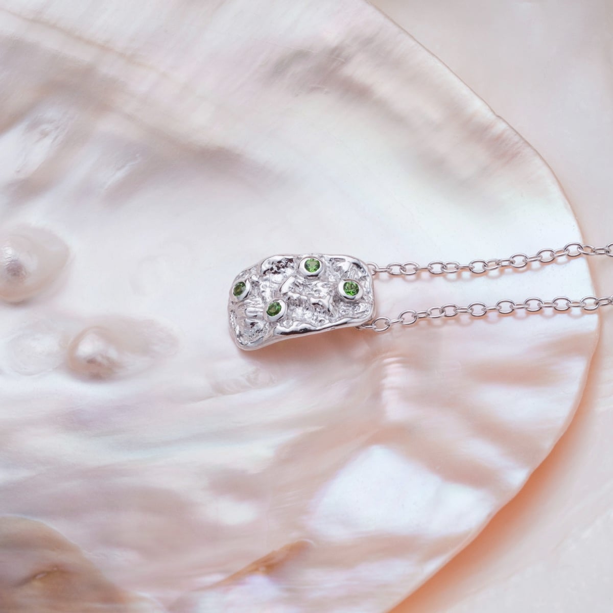 "Silverstream Necklace necklace features a silver design with green diopside accents, shown on a soft, iridescent shell background."