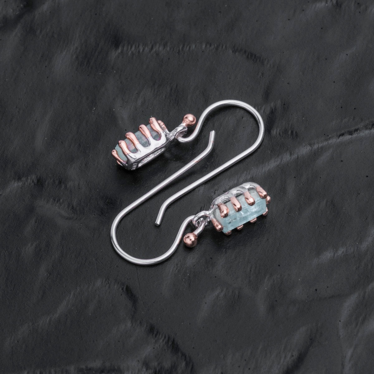 "Silverstream Earrings, elegant earrings with aquamarine stones and rosegold accents, on a dark textured surface."
