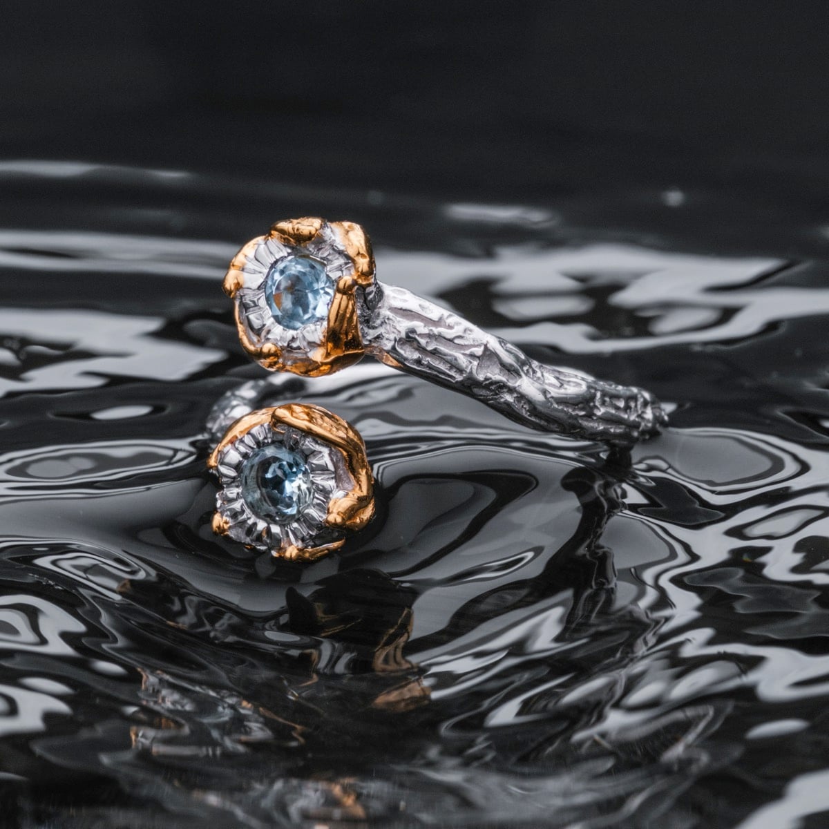 "Silverstream Ring", an elegant ring featuring topaz and gold accents with a textured silver design against a reflective backdrop.