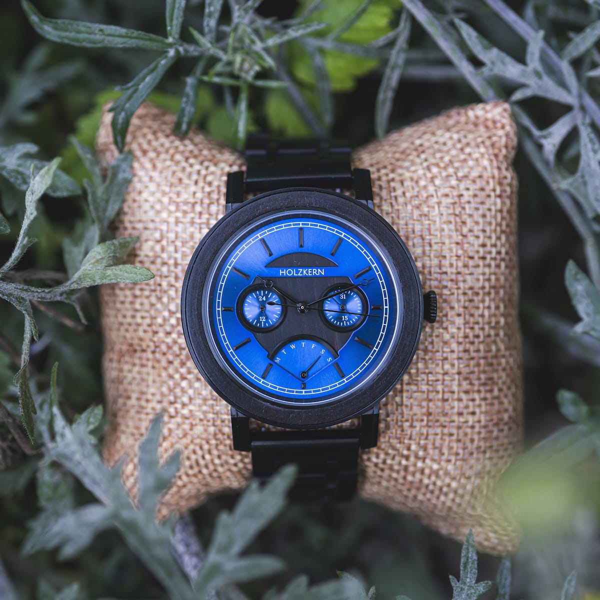 "Belize" watch featuring a blue dial and leadwood accents, displayed on a rustic cushion among greenery.