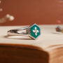 "Spectral Ring" - Un anello in argento e nacre con un gioiello verde esagonale con un design stellato, impostato su uno sfondo caldo.