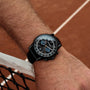 "Rackets watch in black with a sleek, sporty design on a wrist by a tennis court."