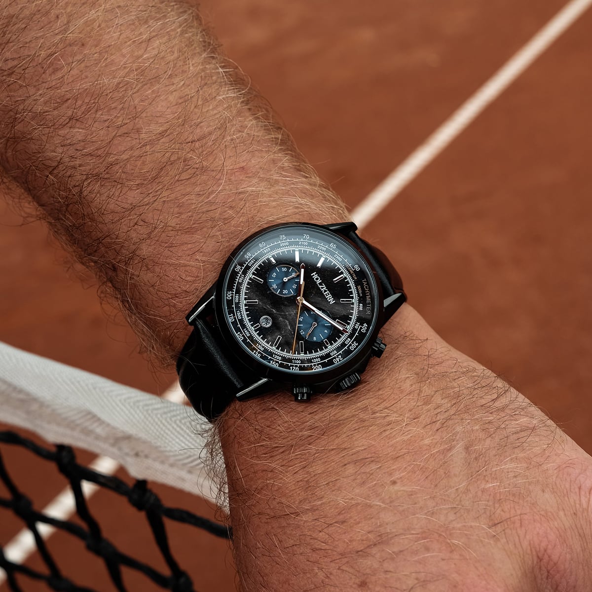 "Rackets watch in black with a sleek, sporty design on a wrist by a tennis court."