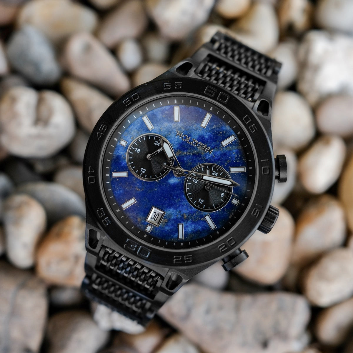 Orologio "Aoraki" con un volto Lapis Lazuli e un involucro nero, impostato su uno sfondo di ciottoli strutturati.