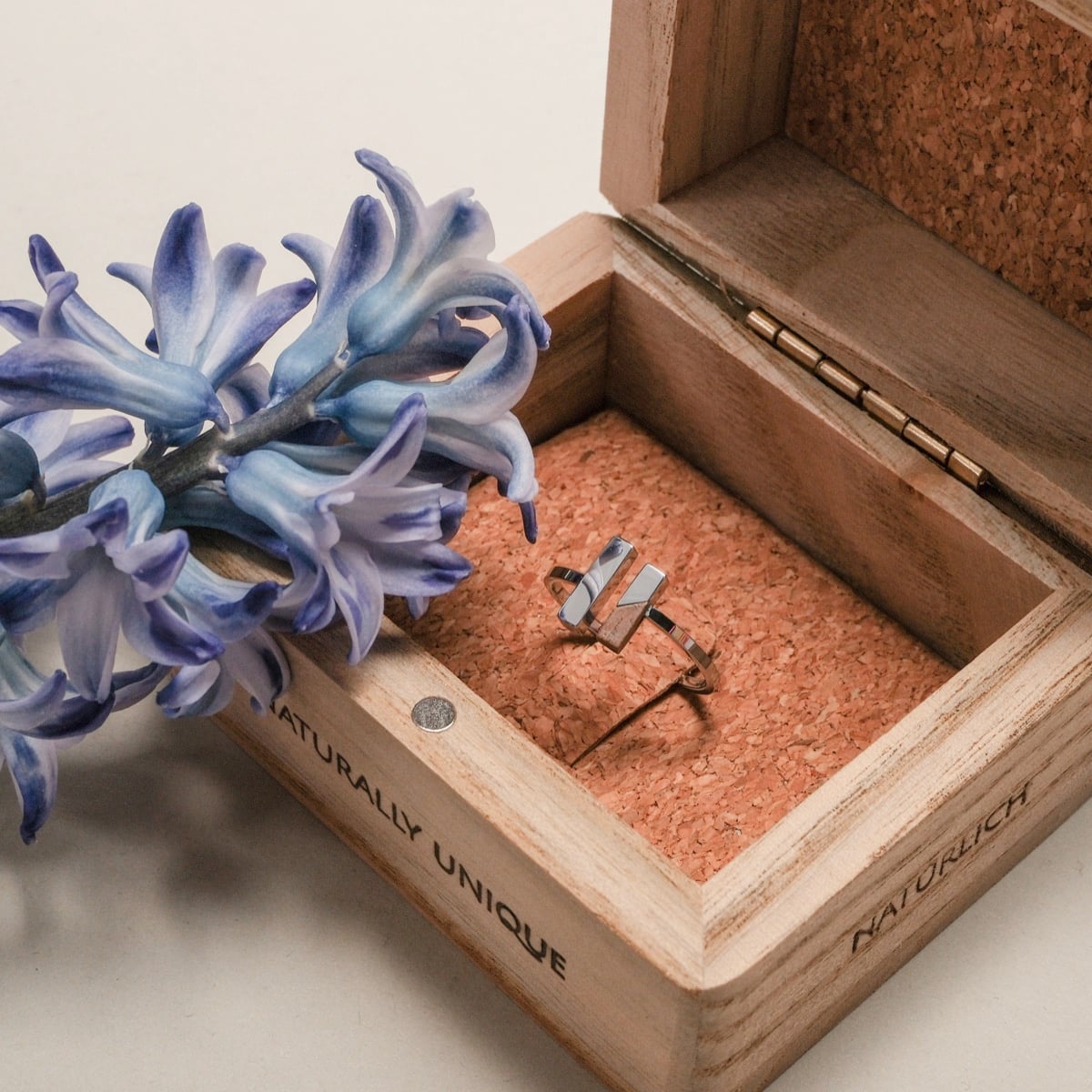 "Campania Ring", a stylish walnut and silver ring in a wooden box, complemented by a blue flower.