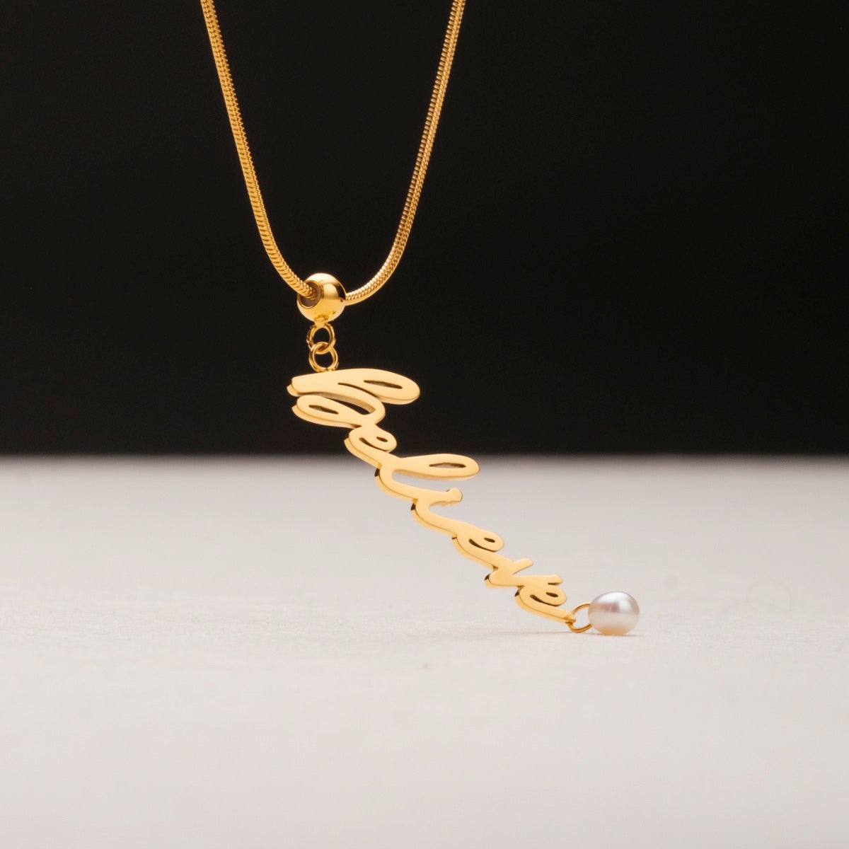 "Believe-Charm", a gold charm necklace featuring a pearl accent, elegant cursive design.