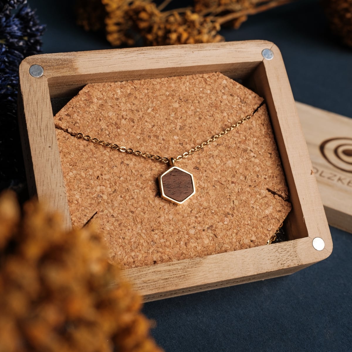 "Collana di legno", una collana di noce esagonale e oro, mostrata in una scatola di legno con fondo di sughero.