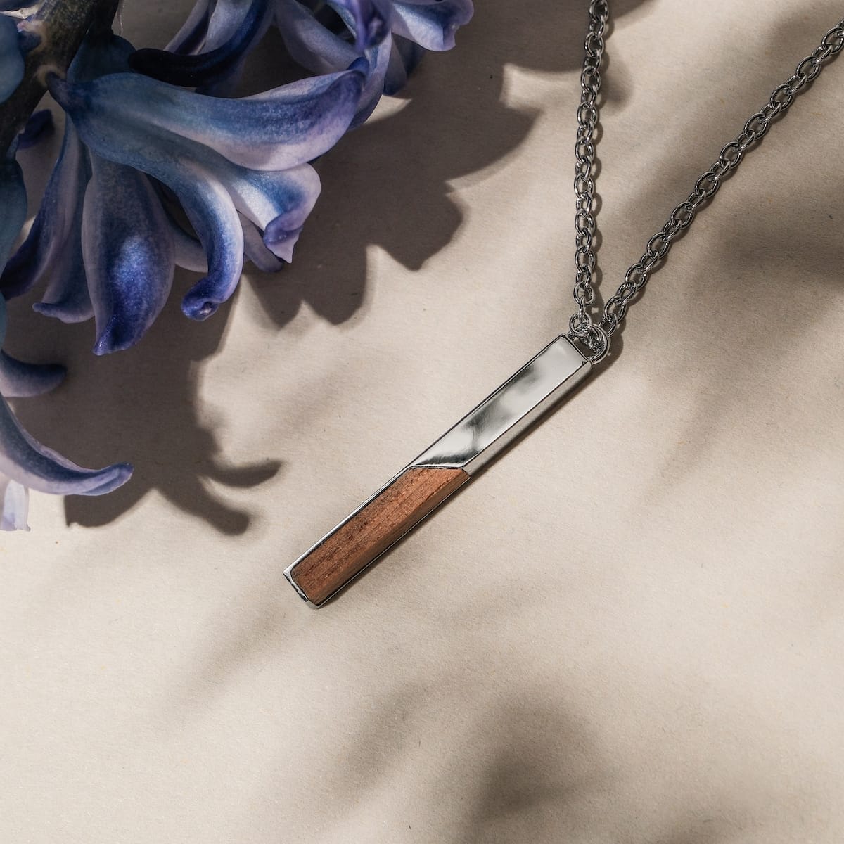 "Campania Necklace" necklace featuring a sleek walnut and silver design on a chain, styled elegantly beside a purple flower.