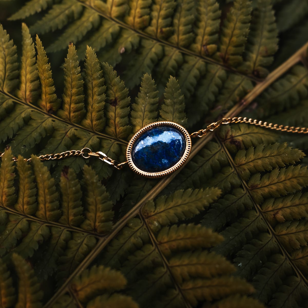 "Bracciale Mirror": Elegante bracciale con un design in oro e lapis lazuli, poggiante su foglie di felce verde vibrante.
