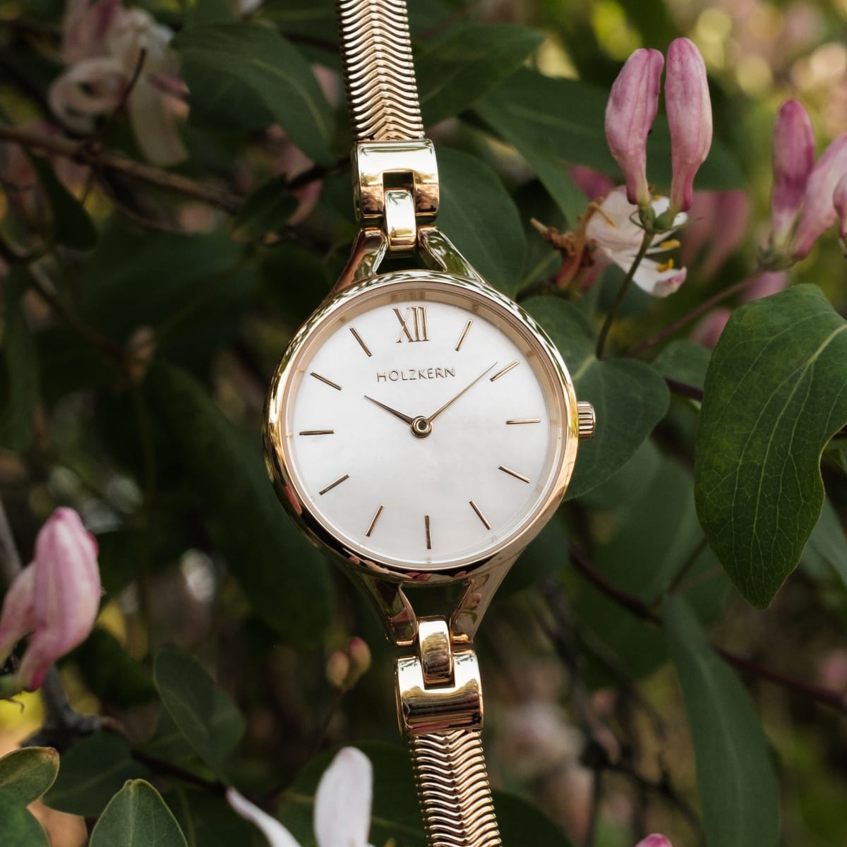 Orologio "Kaliya", nacre e oro, design elegante con stile minimalista in mezzo al verde e fiori rosa.
