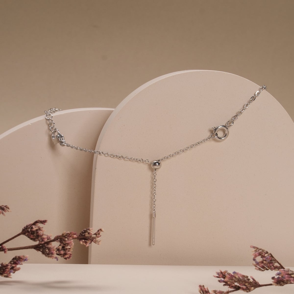 Estenditore di gioielli in argento "Jewelry Extender" su sfondo beige minimalista con fiori decorativi.