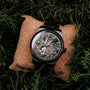 "Adrenin watch, featuring a Cat's Eye style and walnut accents, showcased on a cushion surrounded by greenery."