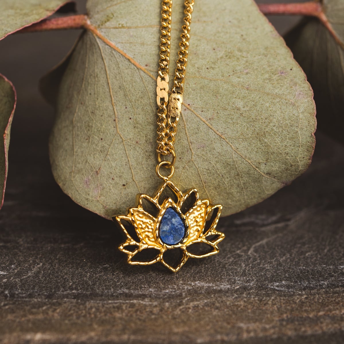 Collana "Balance" con un design di loto in oro con un centro lapis lazuli, impostato contro le foglie naturali.