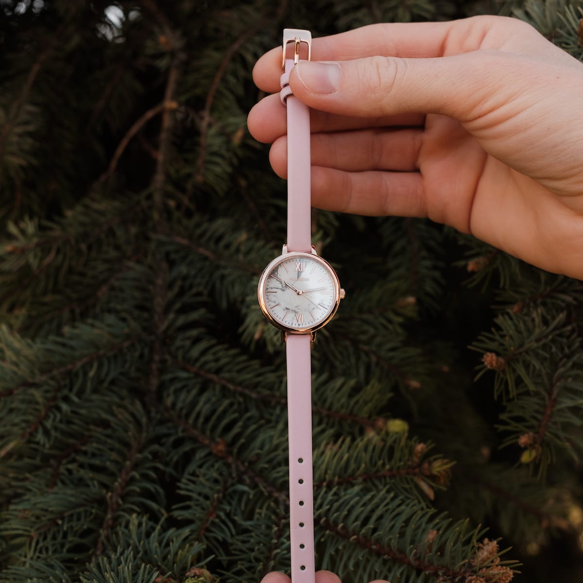 "Alva", a stylish watch with a marble face and rose gold accents, featuring a pink strap held against a pine background.