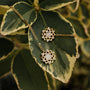 "Composition Gem Earrings": Elegant gold earrings with moonstone detailing, set against variegated foliage.