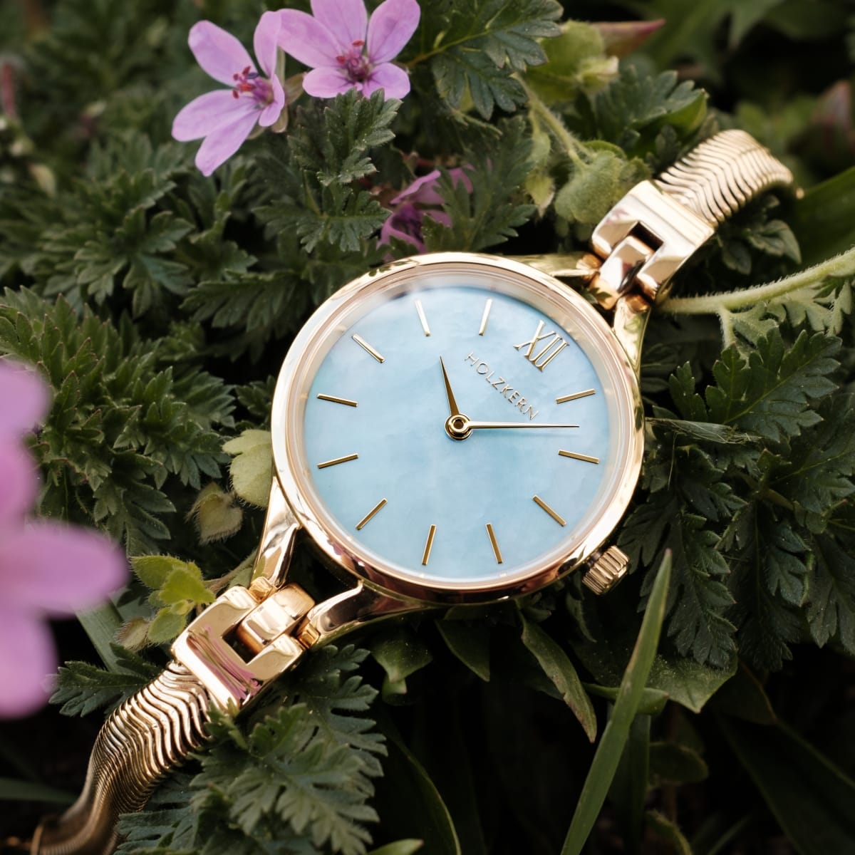 "Shesha" orologio con quadrante nacre e accenti d'oro, che mostra un design elegante e sofisticato tra fogliame verde e fiori viola.