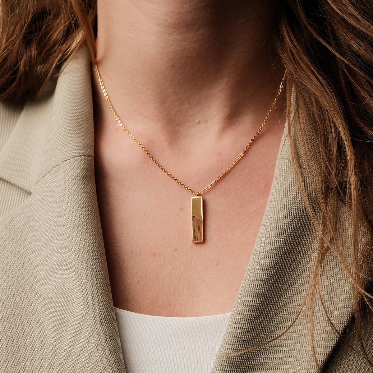 "Ephemera Necklace" necklace, gold chain with a sleek rectangular pendant combining gold and koa wood, worn elegantly on a neutral blazer.