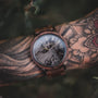 Orologio "Leonardo" con un design in noce e marmo su un polso tatuato, che evidenzia la sua eleganza rustica.