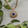 Una collana a ciondolo "Suncatcher" con un design a raggi ultravioletti con lapis lazuli e accenti d'oro, circondata da foglie verdi.