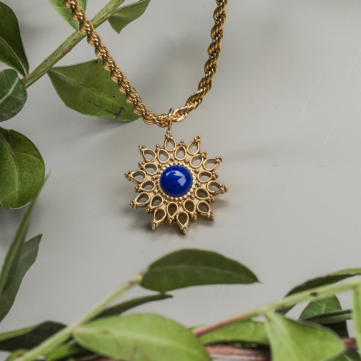 Una collana a ciondolo "Suncatcher" con un design a raggi ultravioletti con lapis lazuli e accenti d'oro, circondata da foglie verdi.
