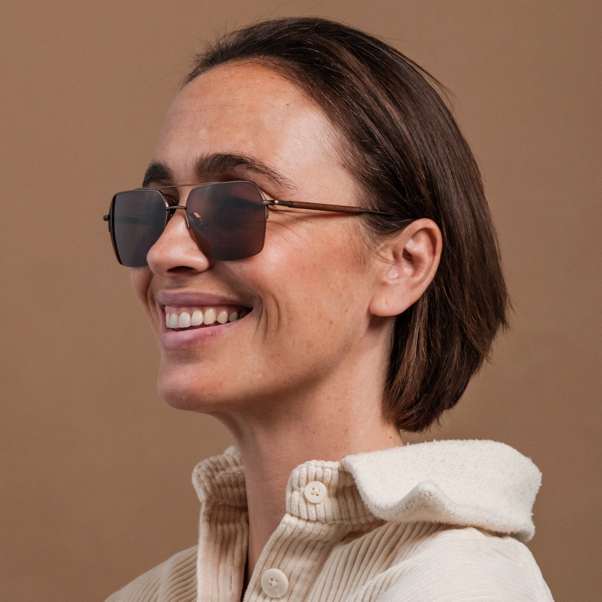 "The Visionary" sunglasses featuring a walnut and bronze style worn by a smiling person in a cozy cream sweater.