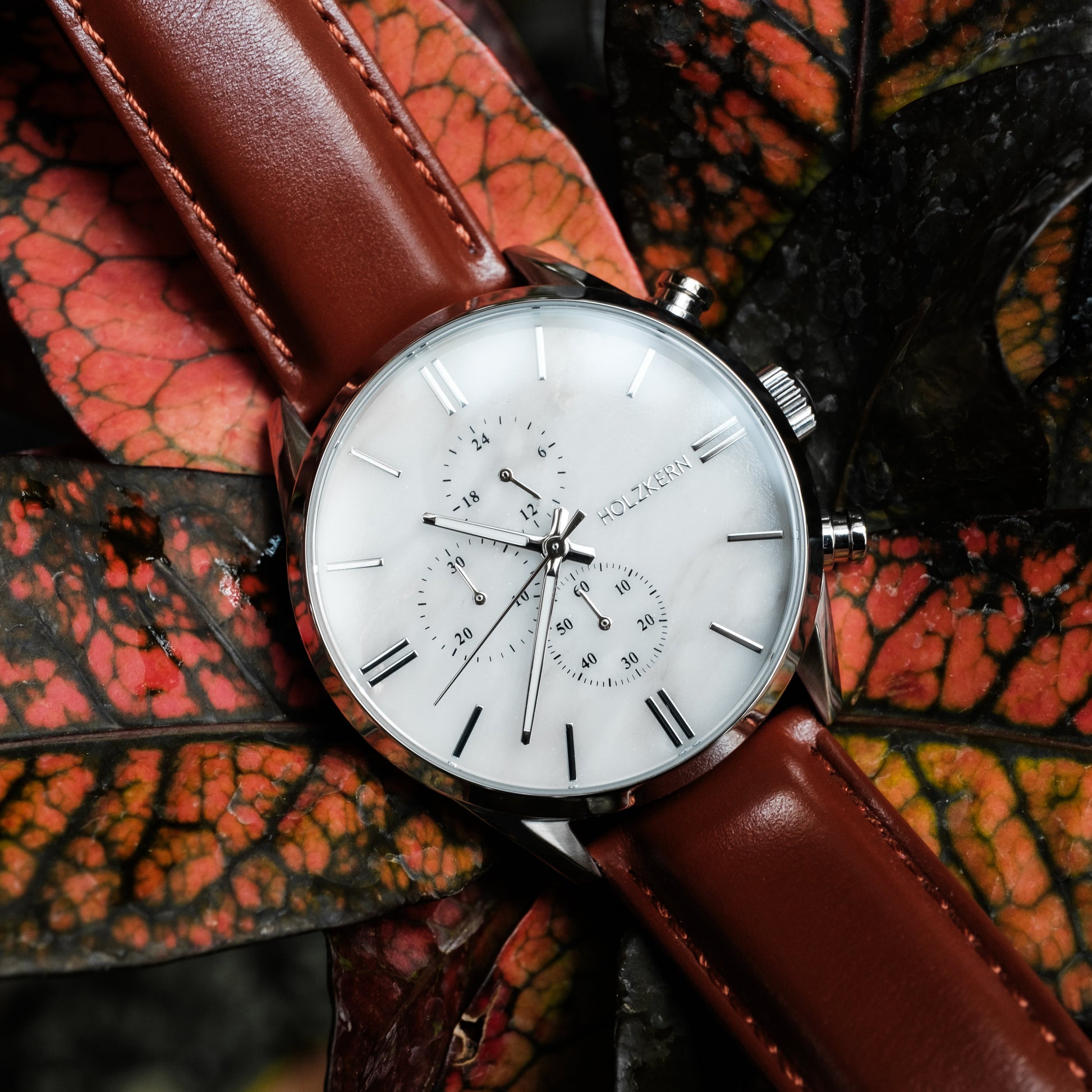Orologio "Melodical" con un viso in marmo, accenti in argento e cinturino in pelle marrone su fondo fogliato strutturato.