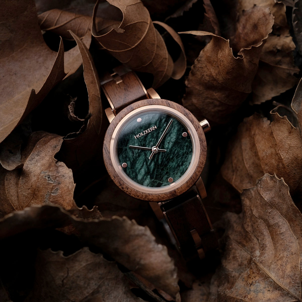 Orologio "Euterpe" con cassa in legno di noce e quadrante in marmo verde, circondato da foglie autunnali.